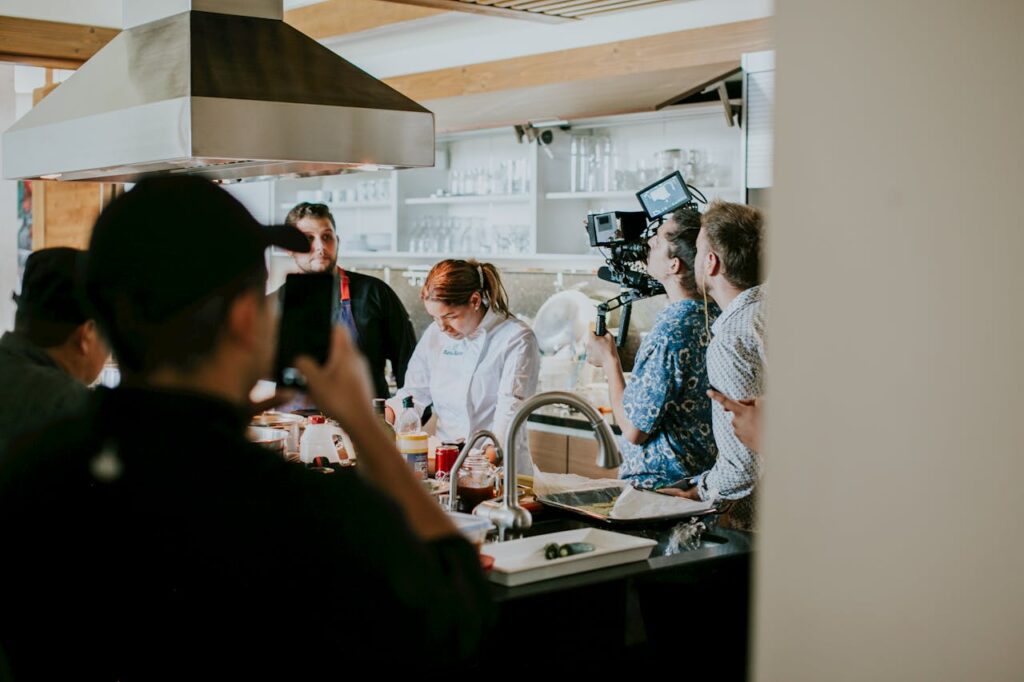 Behind the scenes view of film crew capturing a cooking show in a professional kitchen setting.